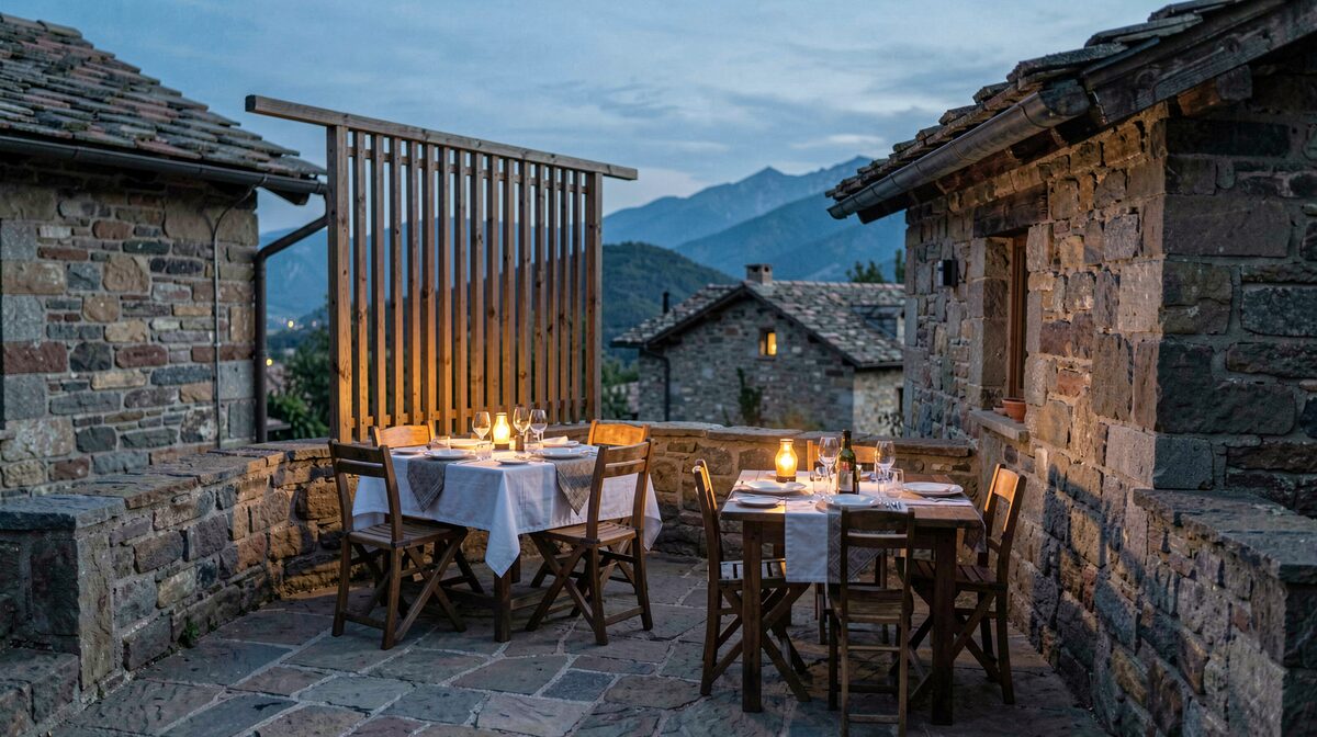A small stone terrace at dusk in a mountain village, with two tables set for dinner, a slatted wooden screen on the windward side, and warm low lamps on each table.